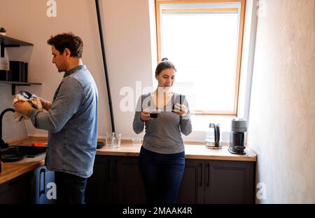 Mari attentionné aidant sa femme avec des plats dans la cuisine. Homme souriant nettoyant les assiettes pendant que son conjoint boit du café. Couple heureux partageant domestiti Banque D'Images