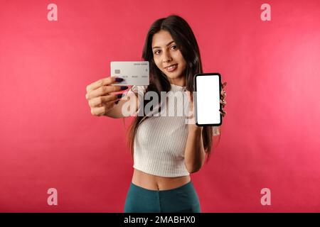 Belle femme brunette portant la récolte blanche striée isolée sur fond rouge recommander boutique, montrant l'écran vide de smartphone et le crédit en plastique Banque D'Images