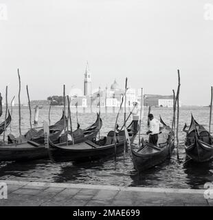 1950s, des rames ou des gondoles vénitiennes historiques et traditionnelles amarrées dans la lagune vénitienne, une baie fermée sur la mer Adriatique, en Italie. Deux gondoliers sont debout dans leurs bateaux. Les bateaux à fond plat sont utilisés pour les promenades autour des îles de Venise, une mosaïque de nombreuses îles, avec la célèbre ville historique de Venise vu au loin. Capitale de la Vénétie, la ville fait partie de l'Italie en 1866. Banque D'Images