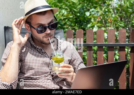 Beau homme avec lunettes de soleil et chapeau de paille boire de l'eau douce avec de la chaux tout en étant assis sur la terrasse en été à l'aide de son ordinateur portable. Banque D'Images