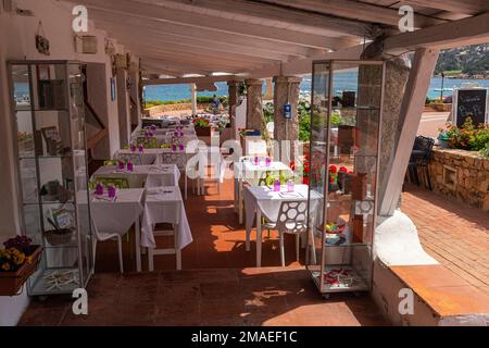 Tables de repas de restaurant à l'ombre d'une marquise en bois avec vue sur la rue et la mer dans la zone commerçante du bijou en bord de mer à Baia Sardaigne. Banque D'Images