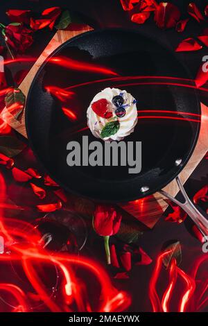 poêle à frire noire avec un gâteau pavlova et des pétales de rose rouges dispersés sur un panneau en bois avec un fond noir avec un verre de vin rouge et des bougies allumées Banque D'Images