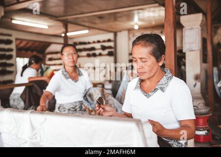 Bali - Indonésie - 10.22.2015: Les femmes indonésiennes âgées dessinant du batik sur du tissu blanc dans un atelier touristique Banque D'Images