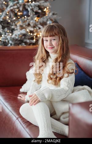 Bonne jeune fille assise sur le canapé près de l'arbre de Noël Banque D'Images