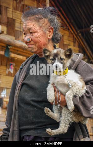 Ziro, Arunachal Pradesh, Inde - 02 24 2009 : Portrait de la vieille femme tribale d'Apatani avec des tatouages faciaux traditionnels et des bouchons de nez portant le chien de chiot Banque D'Images