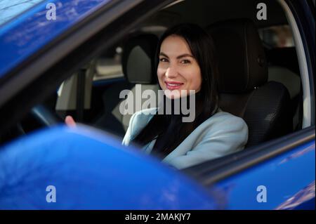 Belle femme européenne d'âge moyen en tenue habillée, voiture de conduite et souriant un magnifique sourire en train de regarder un appareil photo Banque D'Images