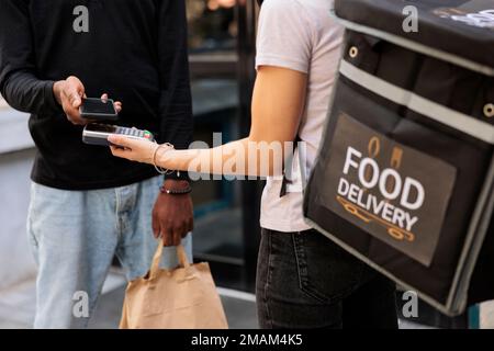 Client effectuant le paiement mobile sans contact pour la livraison de denrées alimentaires de proximité, technologie nfc. Messagerie tenant le terminal de pos., client payant pour un repas à emporter avec vue rapprochée du smartphone Banque D'Images