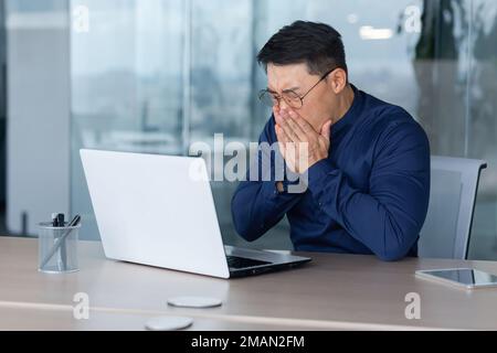 Homme asiatique malade au travail, homme éternuant a de la fièvre et le nez qui coule, homme d'affaires travaillant à l'intérieur du bureau avec un ordinateur portable au travail. Banque D'Images