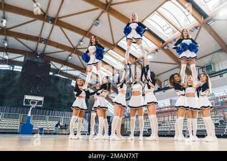 Vue de dessous de l'équipe de meneurs en uniformes bicolores faisant une figure de gymnastique tout en tenant trois membres. Trois membres de l'équipe tiennent des pompons bleus brillants dans leurs mains. Photo de haute qualité Banque D'Images