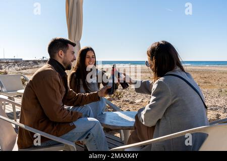 De jeunes amis vêtus de vêtements chauds assis à la table et se trinquer des bouteilles de boissons tout en se reposant le week-end sur la plage Banque D'Images