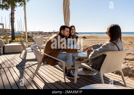 Jeune homme en train de regarder de plus près la main d'amis féminins tout en étant assis à table dans un café extérieur parlant près de la mer le jour ensoleillé Banque D'Images