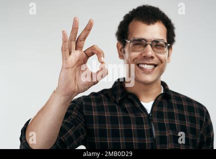 Si personne ne vous l'a dit, je pense que vous faites bien. un beau jeune homme montrant le signe ok tout en se tenant contre un fond gris. Banque D'Images