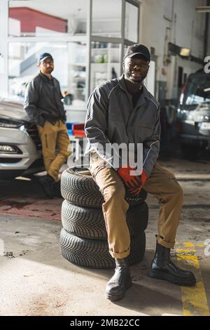 Mécanique automobile dans un atelier de réparation de voiture l'un d'eux assis sur une pile de roues de voiture. Photo de haute qualité Banque D'Images
