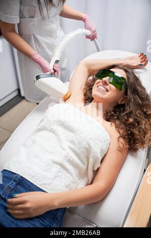 Femme gaie dans des lunettes de protection couchée sur la chaise de jour et souriante tandis que cosmologiste enlever les cheveux de l'aisselle femelle avec dispositif laser. Esthéticienne effectuant une procédure d'épilation au laser dans un salon de beauté. Banque D'Images