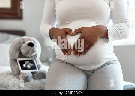 J'adore déjà mon petit paquet. Gros plan d'une femme méconnaissable qui prend une forme de cœur avec ses mains sur son ventre de grossesse à la maison. Banque D'Images