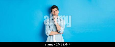Image d'un homme barbu adulte de 25 ans, en pensant à quelque chose, en regardant dans le coin supérieur gauche et en réfléchissant des idées, debout sur fond bleu. Banque D'Images