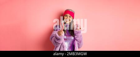 Célébration et fêtes concept. Femme asiatique élégante et âgée s'amusant à faire la fête, mangeant un beignet sucré et se prolongeant la main avec un verre de champagne Banque D'Images