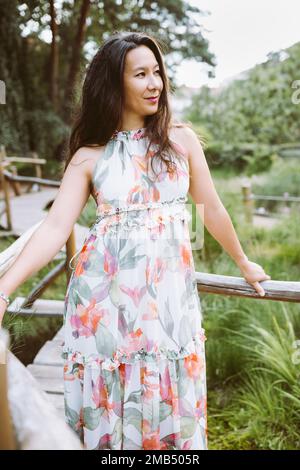 Belle femme japonaise multiraciale heureuse en robe maxi fleurie romantique dans le parc, souriant. Portrait en extérieur. Lumière du soleil, espace de copie Banque D'Images