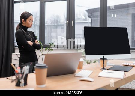 Un designer asiatique croisant les bras près des appareils et du café à aller sur la table en studio, image de stock Banque D'Images