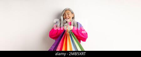 Femme asiatique à la mode, shopaholic, sacs de shopping envieux et souriant joyeuse, l'achat avec des réductions, se tenant sur fond blanc Banque D'Images