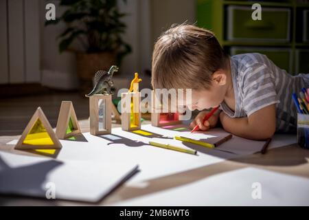 Un petit garçon mignon tire sur le sol de sa chambre. Cercles sur une feuille blanche les ombres des dinosaures projettent de la lumière du soleil. Banque D'Images