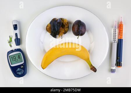 Banane et prunes écrasées sous la forme d'une émoticône d'horreur sur une plaque blanche et des stylos à seringue d'insuline et un glucomètre comme coutellerie sur une table blanche Banque D'Images