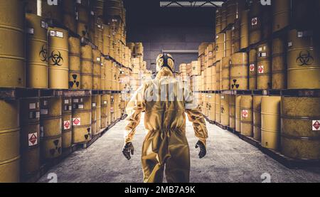 Homme en costume de protection jaune et masque à gaz, entrepôt plein de barils de métal jaune avec des symboles de danger. Banque D'Images