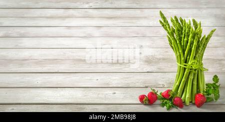 Bouquet d'asperges vertes crues décorées de fraises fraîches sur fond de bois clair et large comme espace de copie Banque D'Images