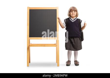 Petite fille dans un uniforme d'école tenant une craie et debout à côté d'un tableau noir d'école isolé sur fond blanc Banque D'Images