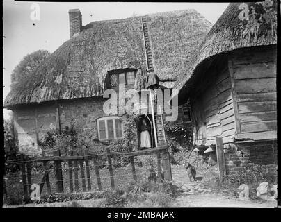 Wootton Rivers, Wiltshire, 1923. Une thatcher travaillant sur le toit d'un chalet non identifié dans les rivières Wootton alors qu'une femme se tient à la porte. Dans le jardin de la maison est un grenier de chaume. La photo semble avoir été prise dans les rivières Wootton, mais l'emplacement exact du chalet est incertain. Banque D'Images