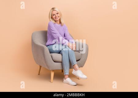Photo pleine grandeur de gaie positive femme habillé violet chandail jeans s'asseoir sur les mains de fauteuil sur les genoux isolés sur fond beige couleur Banque D'Images