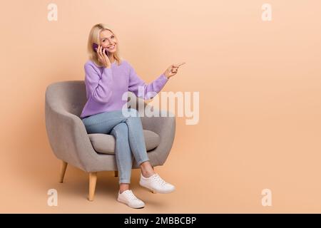 Photo pleine grandeur d'une femme gentille vêtue chandail violet jeans sur le fauteuil parlant en dirigeant l'espace vide isolé sur fond beige couleur Banque D'Images