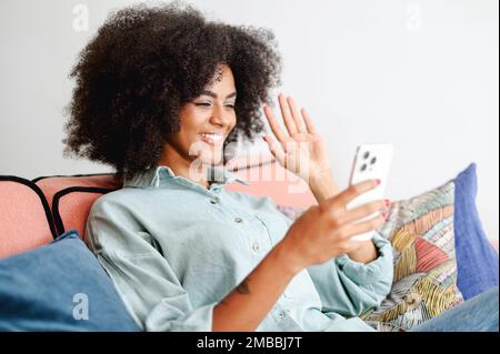 Positive jeune femme noire parlant en ligne sur le téléphone, appel vidéo à la famille ou à l'ami, salutation, dame gaie avec les cheveux d'afro regarde la webcam et la main de la main Banque D'Images
