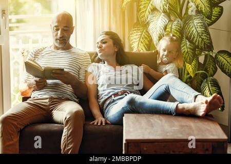 Papa lit à sa fille et à sa mère, petite famille sur le canapé, enveloppé de lumière chaude dans le salon, style de vie familial confortable Banque D'Images