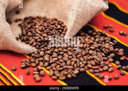 Grains de café torréfiés ou graines dans un sac de jute et éparpillés sur une nappe avec des décorations africaines, gros plan Banque D'Images