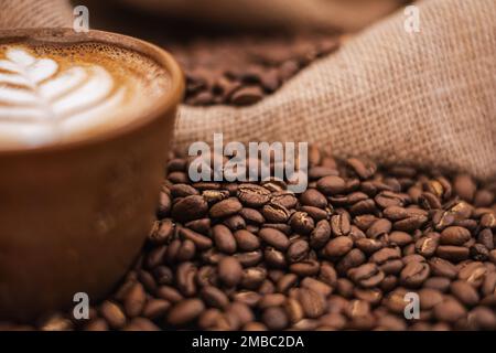 Cappuccino dans une tasse en céramique avec fleur ou plante tirée sur la mousse et torréfié grains ou graines éparpillés sur la table et dans un sac de jute Banque D'Images