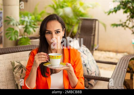 Goiania, Goiás, Brésil – 10 janvier 2023 : une jeune femme, prenant le thé, assise dans une chaise à expression calme, sur le porche de sa maison. Demi-corps Banque D'Images