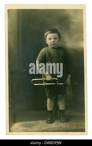 Original charmant début 1900 carte postale de petit garçon mignon avec maison-fait étain jouet trombone, instrument de musique, peut-être un accessoire de studio ou jouet préféré. Le joli petit garçon porte des bottes à lacets, un pull à la main et un short assorti. Vers 1910, 1920, Royaume-Uni Banque D'Images