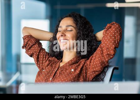Gros plan sur pjoto. Une jeune femme d'affaires latino-américaine est assise au bureau à une table avec un ordinateur portable, les mains derrière sa tête, les yeux fermés, se reposant, souriant. Banque D'Images