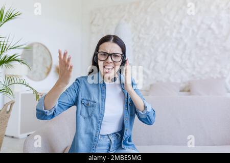 Jeune femme en colère assise à la maison sur le canapé dans un casque, parlant par un microphone, consultation en ligne. Il regarde la caméra, fait des vagues avec colère sur ses mains, explique. Banque D'Images