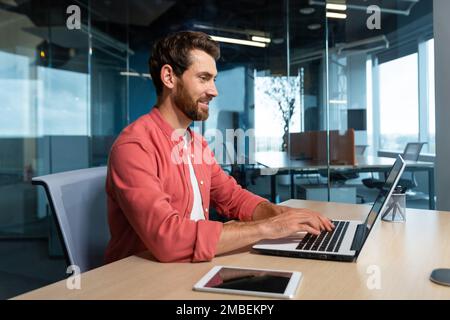 Homme d'affaires réussi en chemise rouge qui travaille avec un ordinateur portable au bureau, homme mûr avec une barbe sur le lieu de travail en tapant sur le clavier souriant satisfait des résultats de travail et des réalisations. Banque D'Images