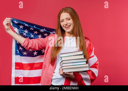 Une étudiante américaine détient une pile de livres universitaires de la bibliothèque universitaire sur fond rose. Bonne fille sourit, elle est heureuse d'obtenir un diplôme aux États-Unis Banque D'Images