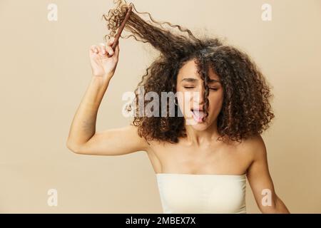 Femme peigne les cheveux bouclés afro avec peigne en bois, produits de coiffage de soins de beauté maison, perte de cheveux après Covid-19, sourire sur fond beige Banque D'Images