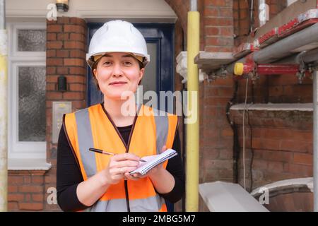 Une femme d'ingénieur civil inspecte et supervise un chantier de construction avec échafaudage, portant un casque et un équipement de protection personnel orange Banque D'Images