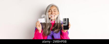 Achats en ligne. Heureuse et ravie asiatique femme senior souriant avec joie, montrant l'écran vide de smartphone et carte en plastique, l'achat dans Internet, blanc Banque D'Images