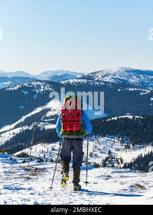 Homme d'âge moyen de l'arrière randonnée pédestre avec des crampons à dos et des bâtons de randonnée en hiver sur une crête de montagne enneigée par temps ensoleillé de près Banque D'Images