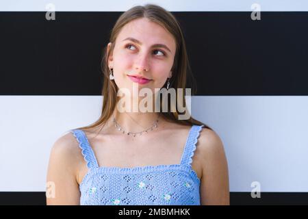 Gros plan d'une adolescente souriante debout devant le mur à rayures noires et blanches et regardant vers le haut et vers la droite Banque D'Images