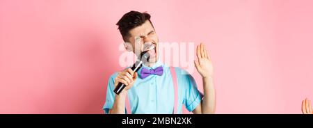 Concept de fête et d'événements festifs. Un gars drôle interprète une chanson, chantant le karaoké dans le microphone avec le visage insouciant, debout dans le nœud papillon et les bretelles sur Banque D'Images