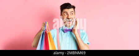 Surpris homme heureux hurlant de joie, regardant de côté le logo et tenant des sacs de shopping, debout sur fond rose Banque D'Images