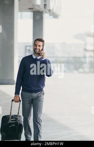 Faire des affaires en déplacement. un beau jeune homme d'affaires en déplacement avec une valise tout en utilisant son téléphone portable pendant la journée. Banque D'Images